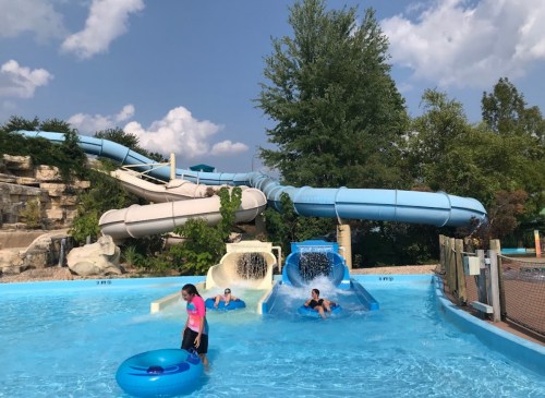 Two water slides with people enjoying the water at a pool, surrounded by trees and a blue sky.