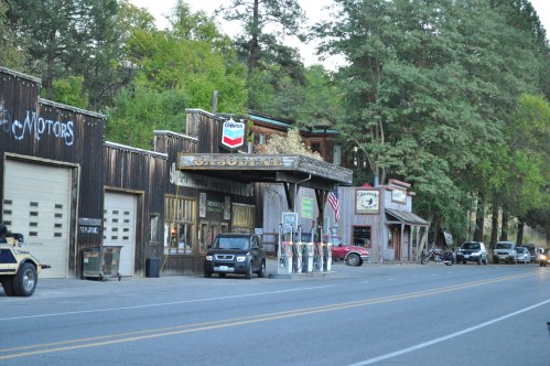 A rustic street scene featuring vintage storefronts, gas pumps, and trees lining the road.