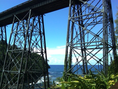 A metal bridge spans a coastal area, with lush greenery and the ocean visible below under a clear blue sky.