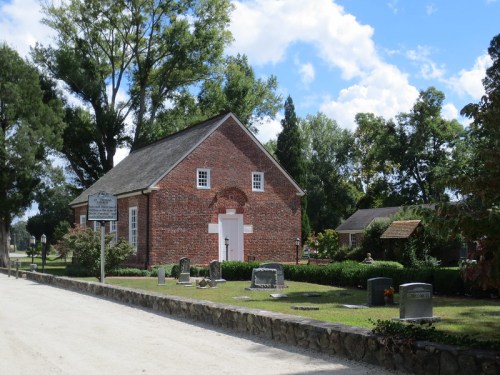 A historic brick building surrounded by trees and gravestones, set along a dirt road under a blue sky with clouds.