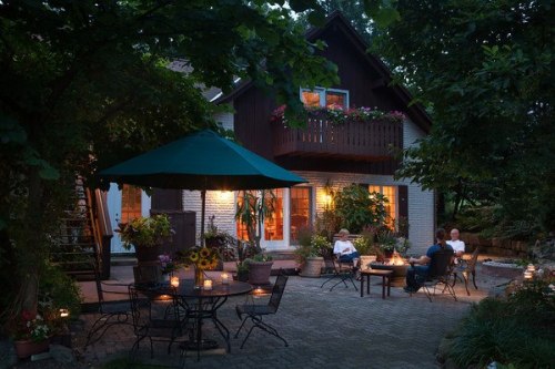 A cozy outdoor gathering with people around tables, candles lit, and a house surrounded by trees in the evening.