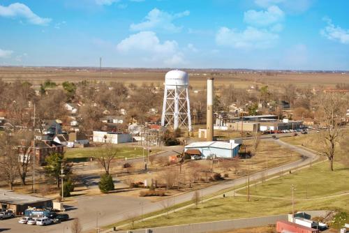 Aerial view of a small town featuring a water tower, industrial buildings, and open fields under a blue sky.