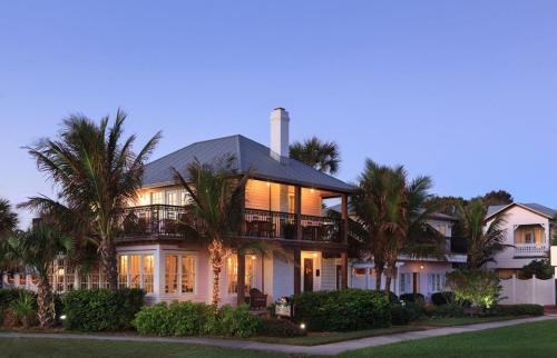 A charming two-story house with a balcony, surrounded by palm trees and lush greenery, at dusk.
