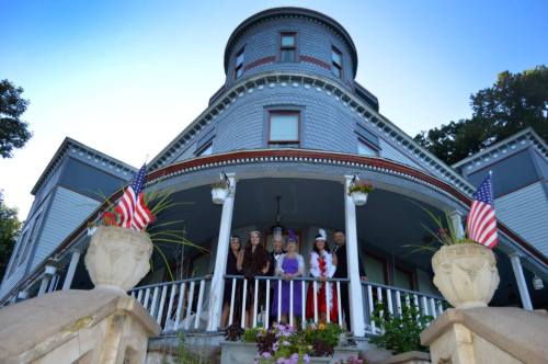 A group of five people in costumes stands on the porch of a large, historic house with American flags and flowers.