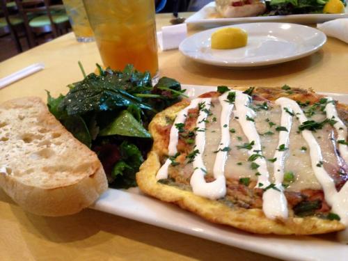A plate with an omelet topped with herbs and sauce, served with a side salad and a slice of bread. A drink is in the background.