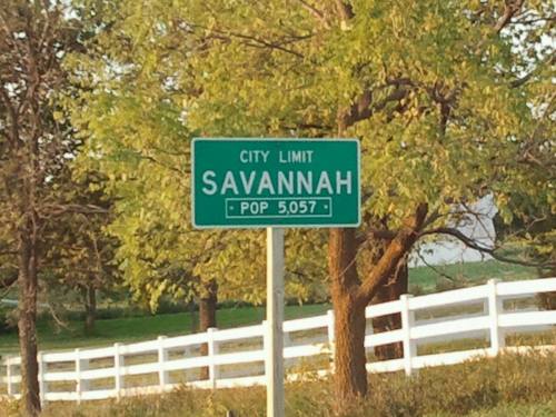 Green city limit sign for Savannah, population 5,057, with trees and a white fence in the background.