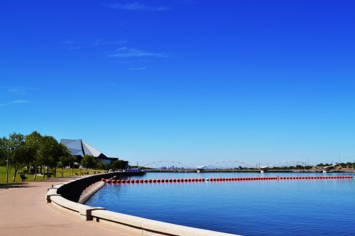A serene lakeside view with a clear blue sky, trees lining the shore, and a bridge in the distance.