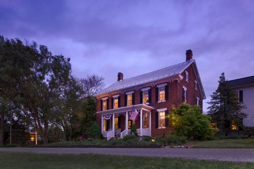 A charming brick house with white trim, illuminated windows, and American flags, set against a twilight sky.