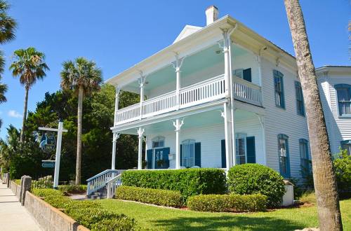 A charming white house with blue shutters and a porch, surrounded by palm trees and greenery under a clear blue sky.