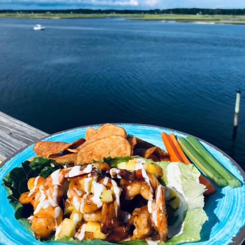 A plate of shrimp with corn on lettuce, drizzled with sauce, served with chips and fresh veggies, by a scenic water view.