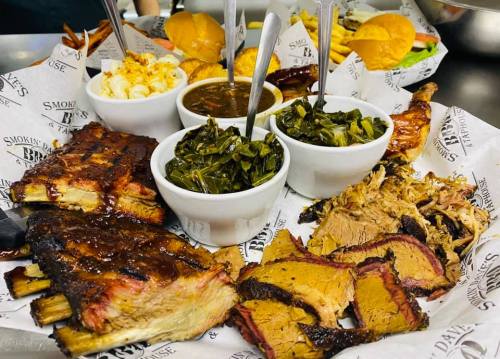 A platter of barbecue featuring ribs, brisket, pulled pork, sides of collard greens, macaroni, and sauces.