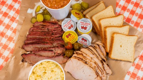 A platter of sliced brisket and turkey, baked beans, pickles, and bread, served with various sauces.