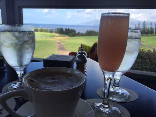 A scenic view of a golf course and ocean, featuring a cup of coffee and two glasses of water and a cocktail on a table.