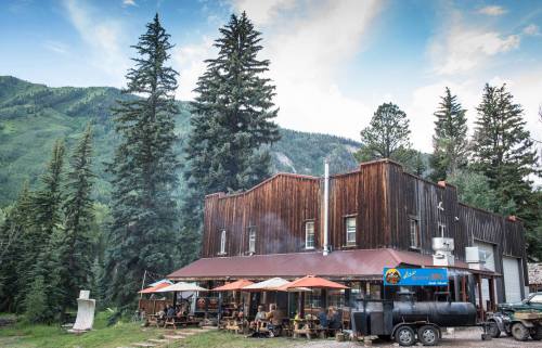 A rustic wooden building with outdoor seating, surrounded by tall trees and mountains under a blue sky.