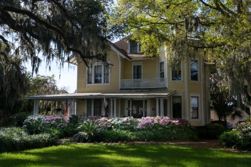 A charming yellow Victorian house surrounded by lush greenery and colorful flowers under a sunny sky.