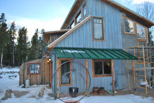 A rustic blue house with a green roof, surrounded by snow and trees, featuring a hanging chair and construction scaffolding.
