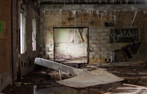 An abandoned room with debris, peeling walls, and a doorway leading to a dimly lit space beyond.