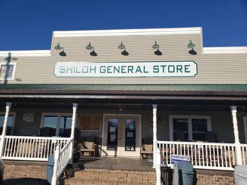 Front view of Shiloh General Store, featuring a green sign and a porch with rocking chairs. Clear blue sky above.