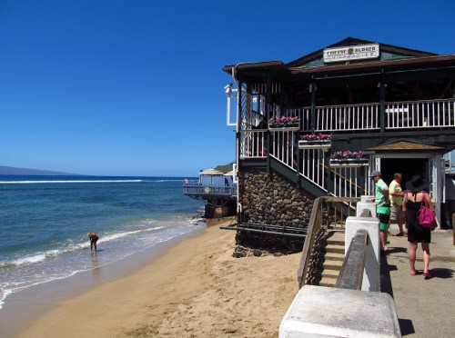 A beachside building with a deck, people walking along the shore, and clear blue skies in the background.