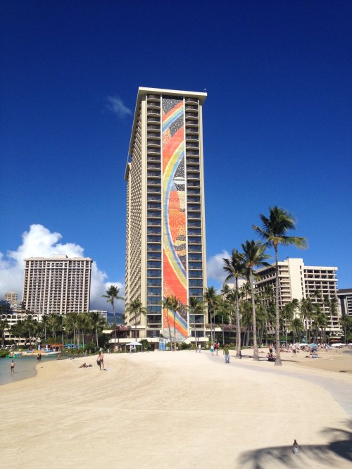 A tall building with a colorful mural stands near a sandy beach, surrounded by palm trees and a clear blue sky.