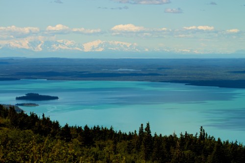 A serene landscape featuring a turquoise lake, distant snow-capped mountains, and lush green forests.