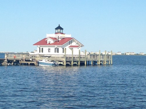 A white lighthouse with a red roof on a pier, surrounded by calm blue water and a clear sky.