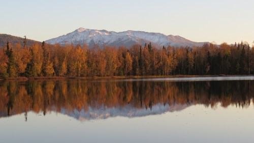 A serene lake reflects autumn trees and snow-capped mountains under a clear sky.