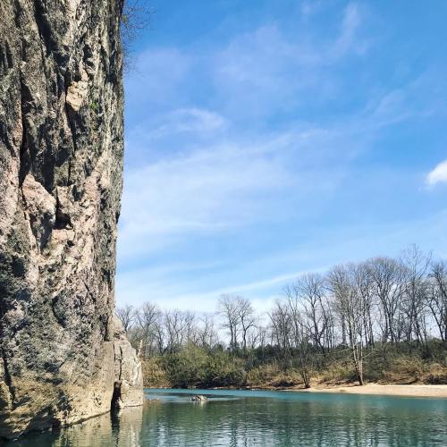 A serene river scene with a rocky cliff on one side and bare trees lining the opposite bank under a blue sky.