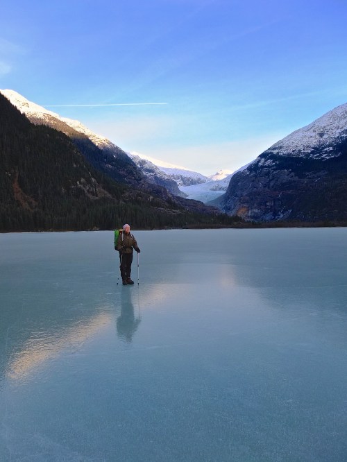 A person stands on a frozen lake surrounded by snow-capped mountains and a clear blue sky.