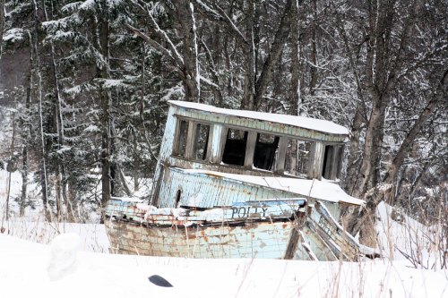 Abandoned, weathered boat named "Polly" partially buried in snow, surrounded by trees in a winter landscape.