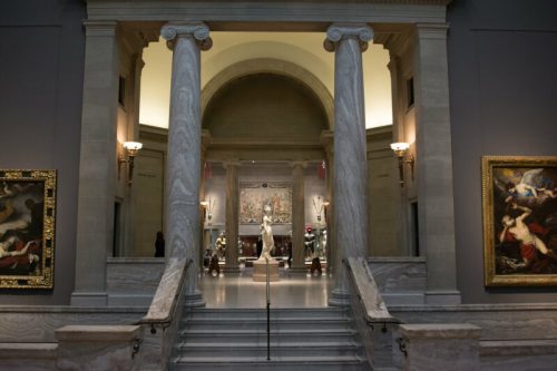 A grand museum entrance with marble columns, stairs, and artwork lining the walls, leading to a central exhibit.