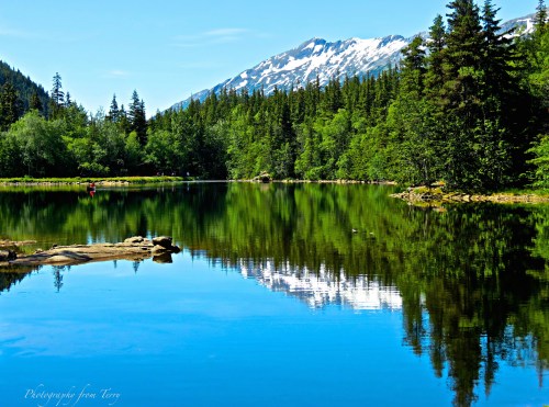 A serene lake reflecting snow-capped mountains and lush green trees under a clear blue sky.