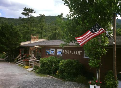 A rustic restaurant building surrounded by greenery, with an American flag waving in front and mountains in the background.