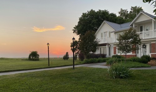 A serene sunset over a grassy field, with a charming house and lampposts in the foreground.