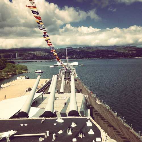 View from a ship's deck featuring large cannons, colorful flags, and a scenic harbor with mountains in the background.