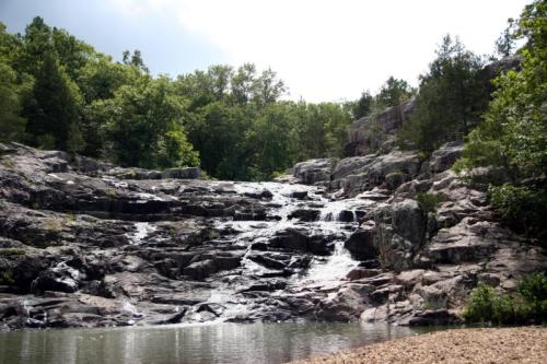 A serene waterfall cascading over rocky terrain, surrounded by lush green trees under a partly cloudy sky.