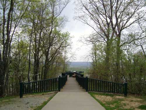 A paved path leads through trees to a scenic overlook with a railing, surrounded by greenery and distant hills.