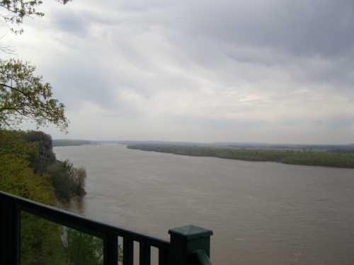 A wide river flows through a lush landscape under a cloudy sky, viewed from a green railing.