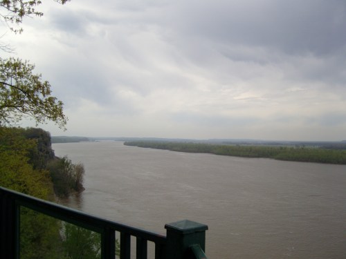 A wide river flows through a lush landscape under a cloudy sky, viewed from a green railing.