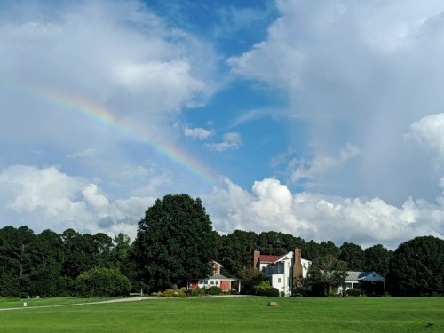 A vibrant rainbow arcs over a green field and houses under a partly cloudy sky.