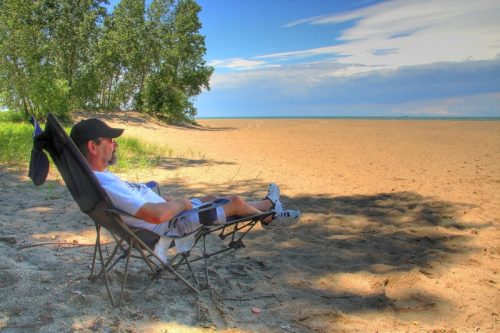 A man relaxes in a chair on a sandy beach, surrounded by trees and a calm lake under a blue sky.