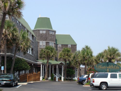 A charming hotel with a green roof, surrounded by palm trees and parked cars in a sunny setting.