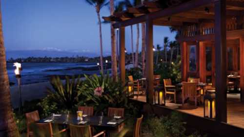 A beachfront restaurant at dusk, surrounded by palm trees and lanterns, with tables set for dining.