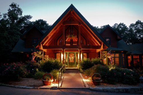 A warmly lit lodge with a large wooden entrance, surrounded by greenery and flower pots, at dusk.
