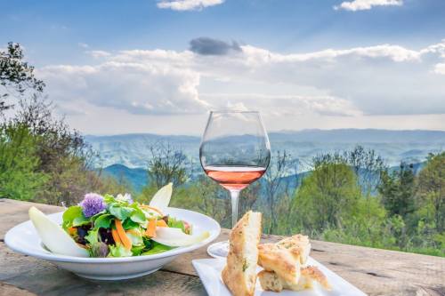 A scenic view of mountains with a plate of salad, bread, and a glass of rosé wine on a wooden table.