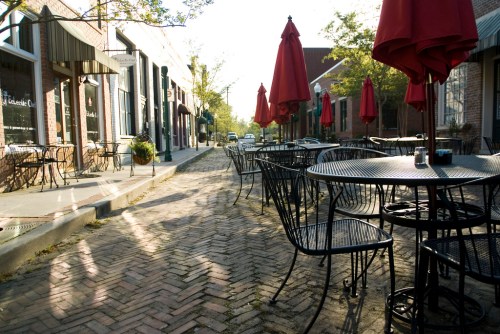 Empty outdoor café with black metal tables and red umbrellas lining a brick-paved street in a quiet neighborhood.