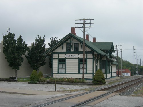 Historic train station in Higginsville, featuring a green roof and white siding, surrounded by trees and power lines.