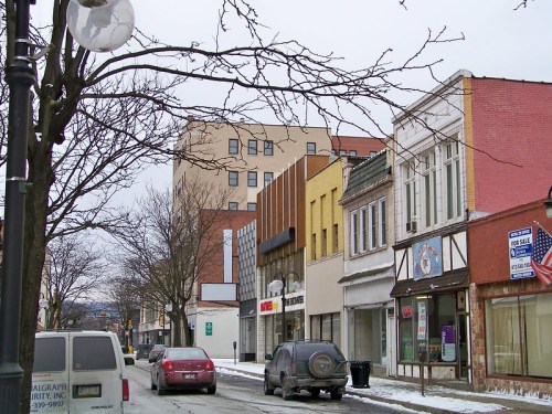 A snowy street scene featuring shops, parked cars, and a mix of modern and historic buildings.