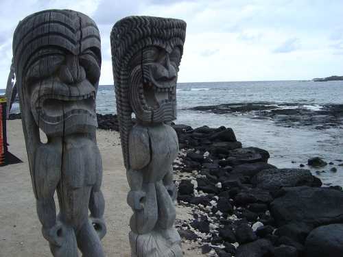 Two wooden tiki statues stand on a beach, overlooking the ocean and rocky shoreline under a cloudy sky.