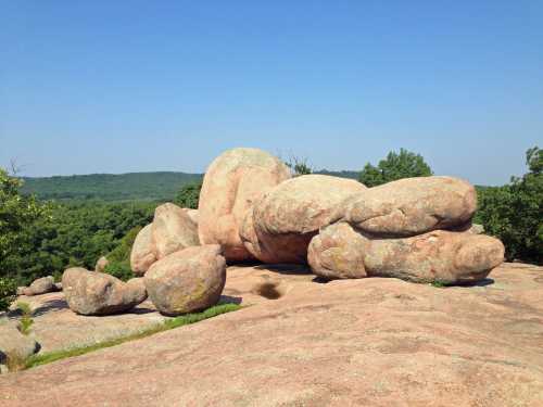 A large, weathered rock formation sits atop a hill, surrounded by green trees and a clear blue sky.